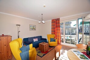 Ein Wohnzimmer mit einer Couch, zwei Sesseln, einem Couchtisch und einem Esstisch mit Stühlen. Ein Fenster bietet Blick auf Gebäude.