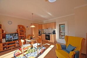 A living room with a television, wooden furniture, a yellow armchair, and a kitchen in the background. - Just 50 meters from the sandy Baltic Sea beach, Wi-Fi, washing machine, accommodates up to 4 people, with a balcony (Glowe)