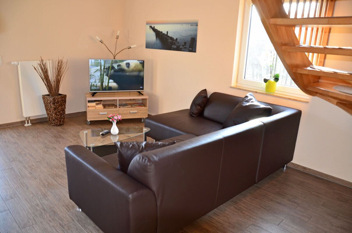Ein modernes Wohnzimmer mit einem Ecksofa, einem Glastisch, einem Fernseher und einem Fenster mit Blick auf eine Holzkonstruktion.