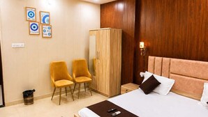 A hotel room with a bed, a wardrobe, two chairs, a small table, and a trash bin. - The Shree Heritage And Rooftop (Bara Banki)