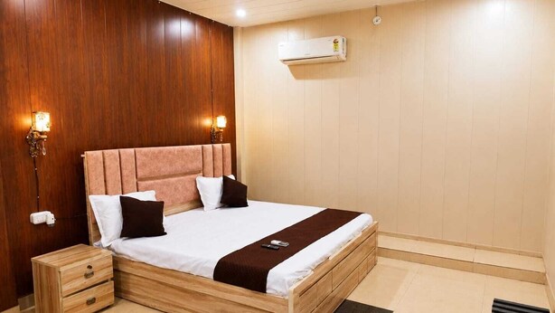 A hotel room with a wooden headboard, a bed with white and brown bedding, a bedside table with drawers, and an air conditioning unit on the wall. - The Shree Heritage And Rooftop (Bara Banki)