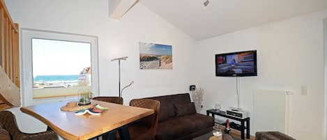 Un salon moderne avec une table à manger en bois, des canapés marron, un téléviseur à écran plat et une vue sur la plage, visible à travers une grande fenêtre.
