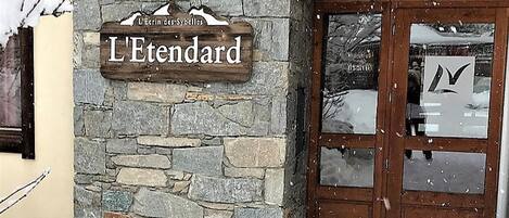 Edifício de pedra com uma placa de madeira que diz 'L'Etendard' e uma porta com painel de vidro.