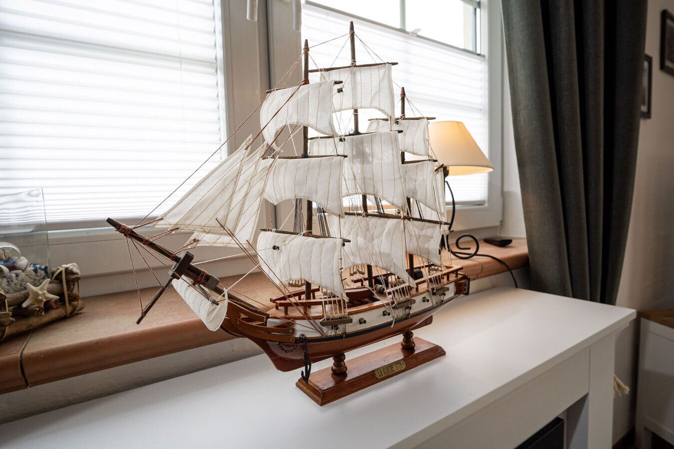 Une maquette de bateau, posée sur un socle en bois, dans une pièce dotée d’une fenêtre et d’une lampe.
