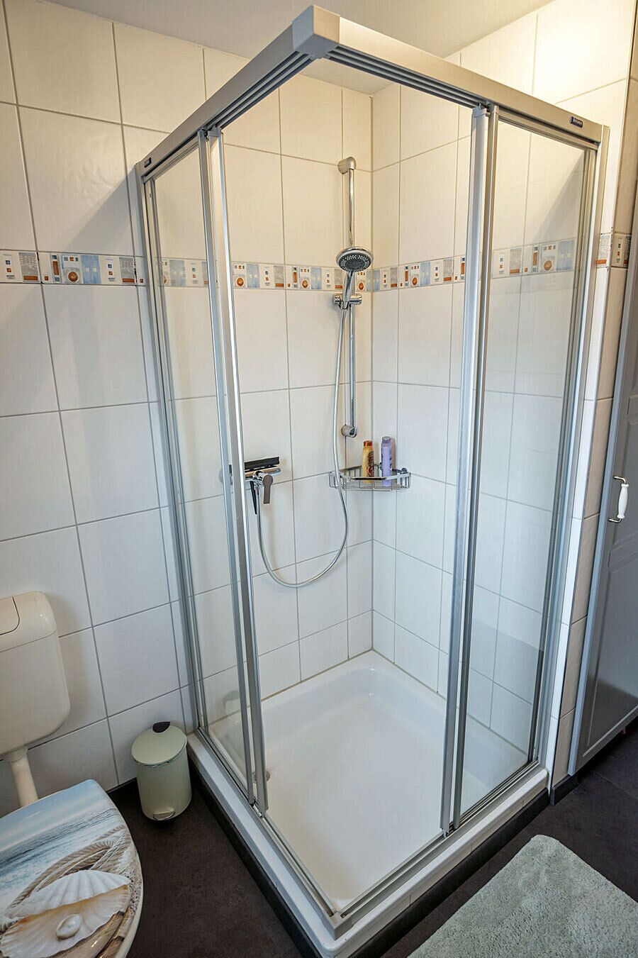 Cabina doccia in vetro con soffione a mano, rubinetto a parete e piccolo ripiano con articoli da toilette.