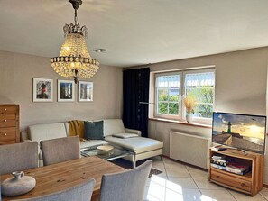 A living room with a sofa, a dining table, a chandelier, a television, and a window with a view of greenery. - 900 meters to the beach, 2 bedrooms, sleeps up to 4, Wi-Fi, pets allowed by arrangement (Breege)