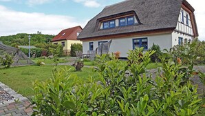 Um edifício tradicional com telhado inclinado, um prolongamento de telhado de colmo e um jardim bem cuidado com diversas plantas.