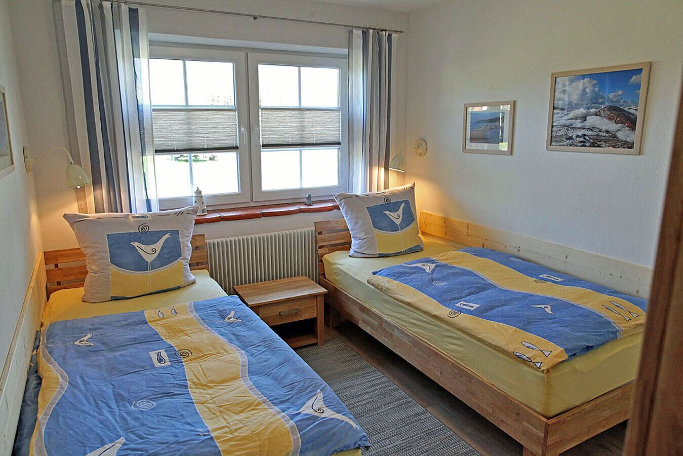 Une chambre avec deux lits, une fenêtre avec des stores, un radiateur et une petite table.