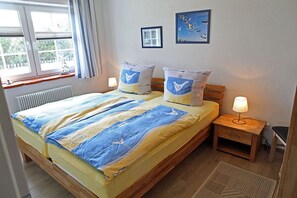 Ein Doppelbett mit gestreifter Bettdecke, zwei Kissen mit Vogelmotiv, ein Nachttisch mit Lampe, ein gerahmtes Bild an der Wand und ein Fenster mit Vorhängen.