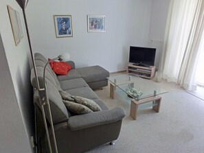 Ein Wohnzimmer mit grauem Sofa, einem Glastisch, einem Flachbildfernseher und gerahmten Kunstwerken an der Wand.