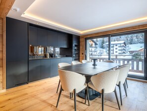 A modern kitchen with dark cabinetry, a dining table with chairs, and a view of a snowy landscape through large windows. - Bright apartment - 10 guests (Megève)