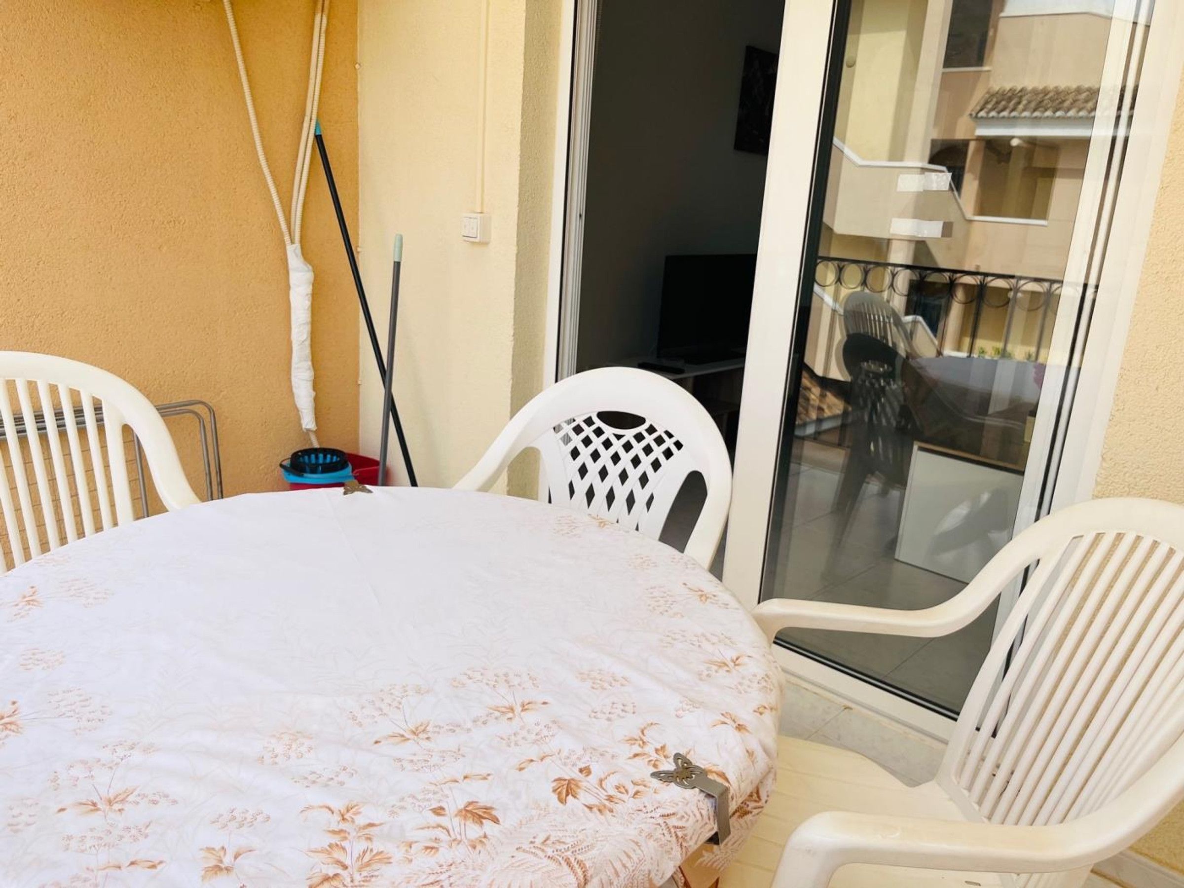 Une pièce avec une table ronde recouverte d’une nappe à motifs floraux, deux chaises en plastique blanches, un balai et un seau.