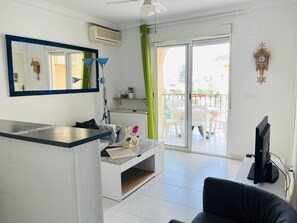 Una sala de estar moderna con un televisor de pantalla plana, un sofá, una mesa de centro y un ventanal corredero que da a un balcón con mobiliario exterior.