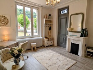 A room with a fireplace, a radiator, a white sofa, a small table with a vase, and a large window with a view of greenery.