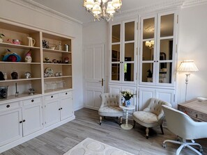 A room with a large white cabinet, two armchairs, a small table with a vase, and a chandelier.