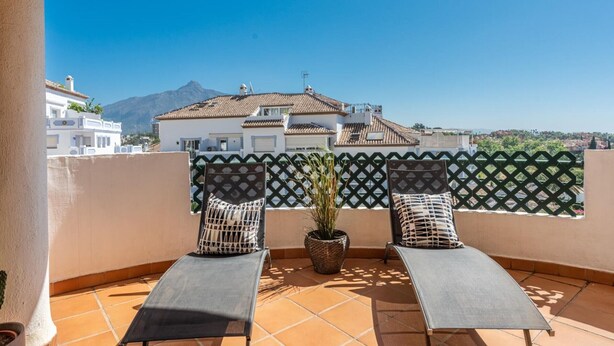 Una terraza con dos sillones, una planta en maceta y vistas a la montaña y edificios.