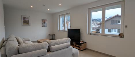 Uma sala de estar moderna com um sofá cinza, uma televisão e uma janela grande com vista para uma área residencial.