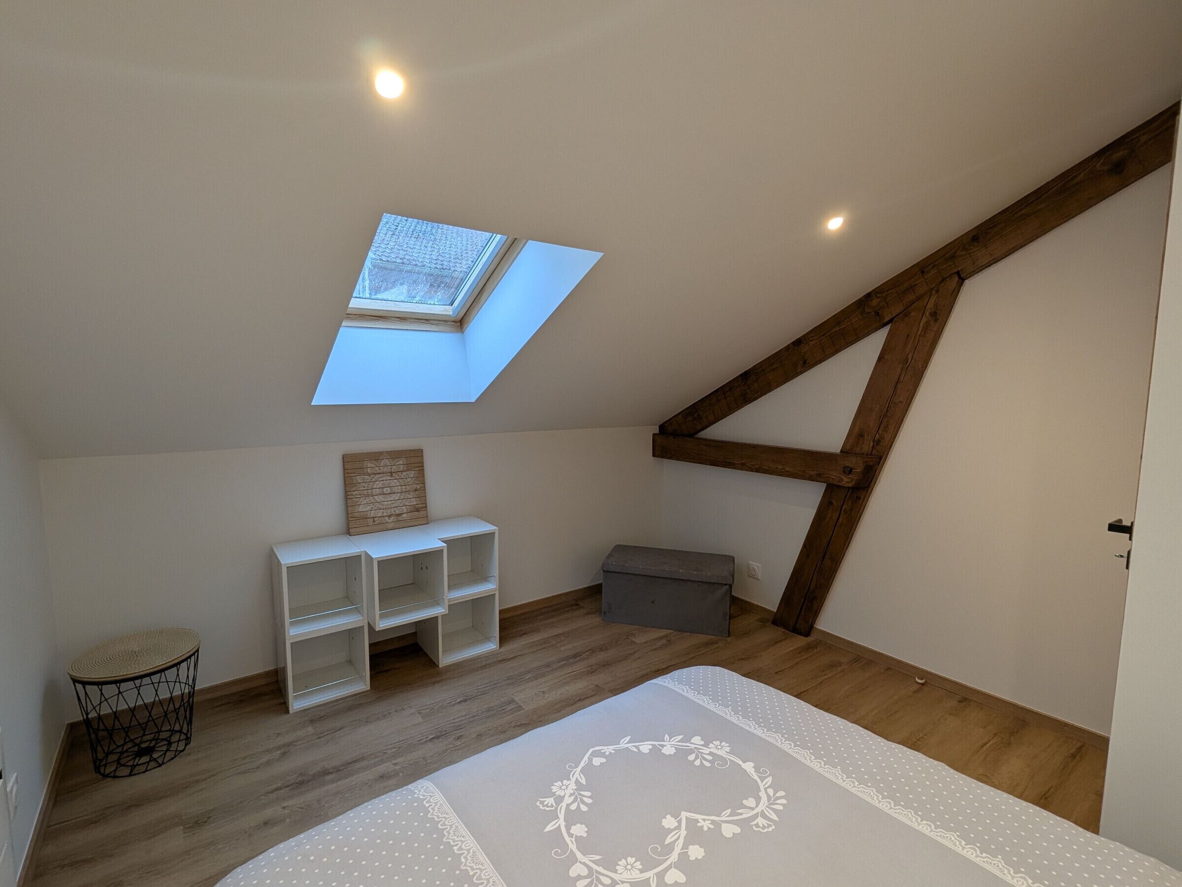 A room with a sloped ceiling, a skylight, wooden beams, a white bench, a small grey storage box, and a patterned rug.