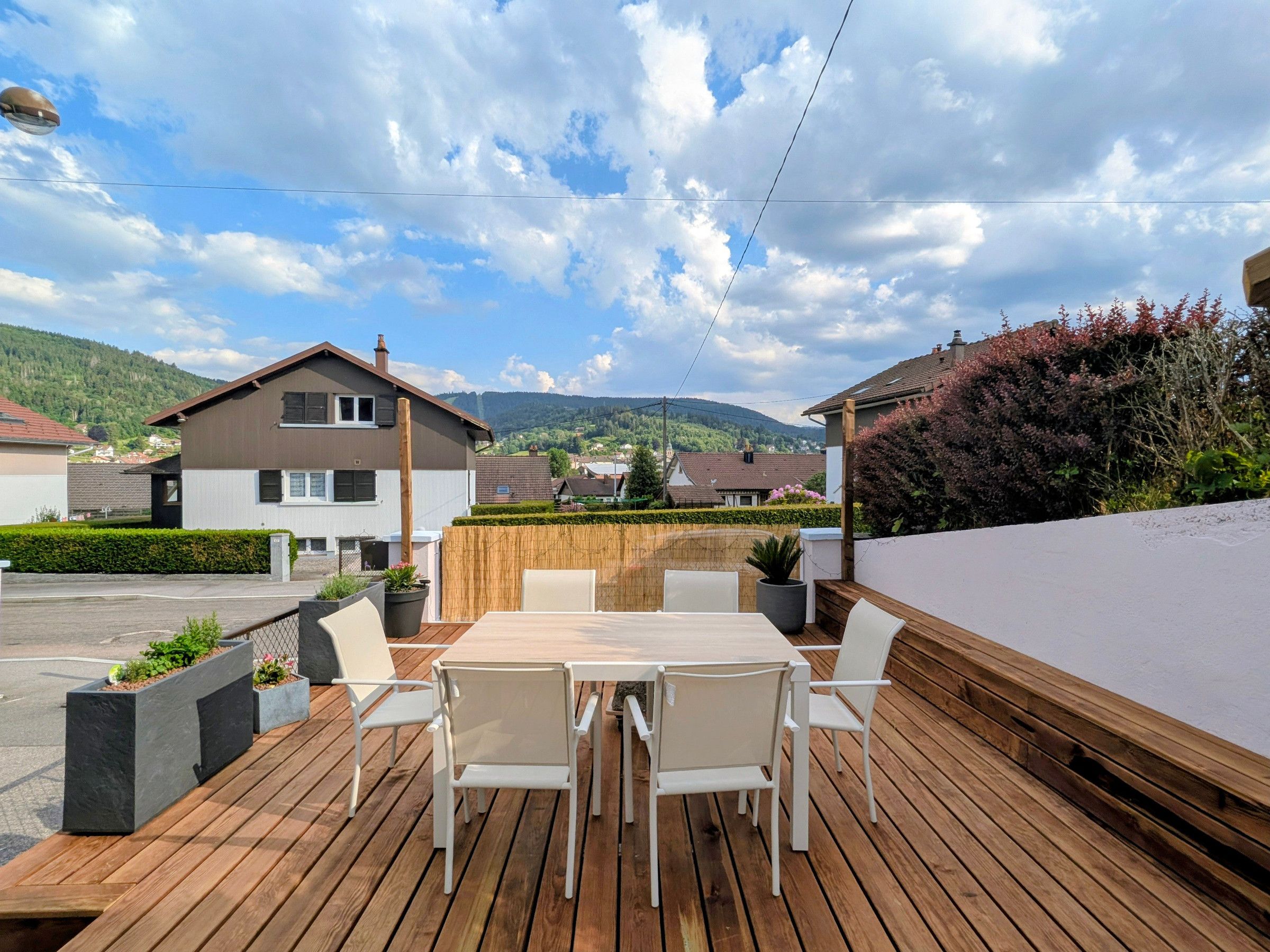 Eine Holzterrasse mit Tisch und Stühlen, umgeben von einer niedrigen Mauer und Kübelpflanzen.