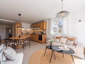Uma cozinha moderna com armários de madeira, uma área de jantar com mesa e cadeiras redondas, uma sala de estar com sofá e mesa de centro, e uma grande janela com cortinas.