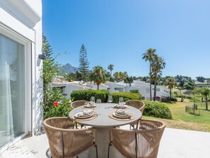 Uma sala de jantar com uma mesa posta para quatro pessoas, rodeada por cadeiras de vime, com vista para uma área residencial com casas e vegetação.