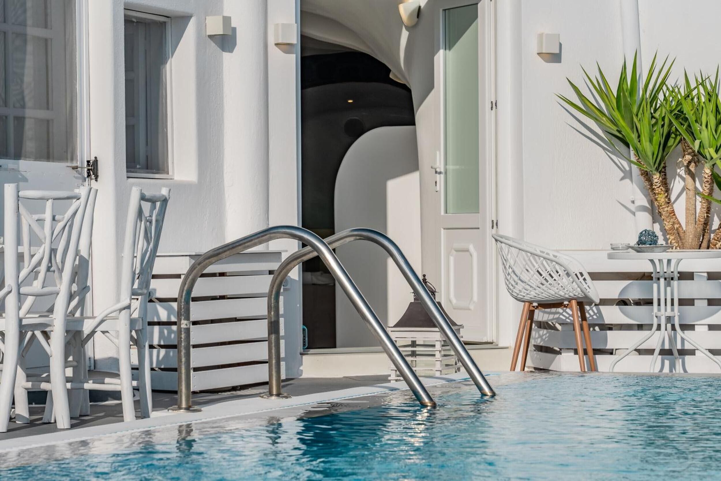 Zona de piscina con una escalera metálica que da al agua, una silla blanca y una mesa con una planta al lado.