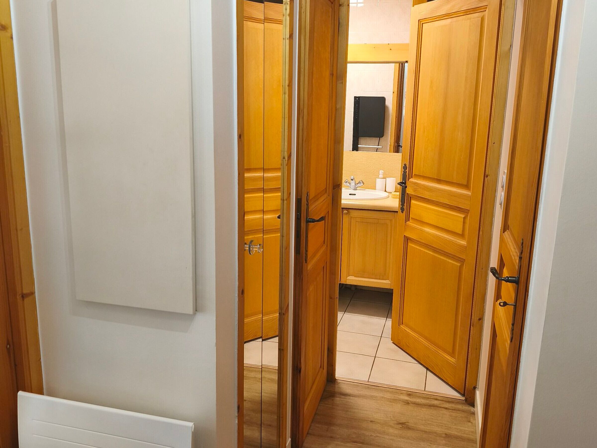 A narrow hallway with wooden doors leading to a bathroom with a sink and mirror.