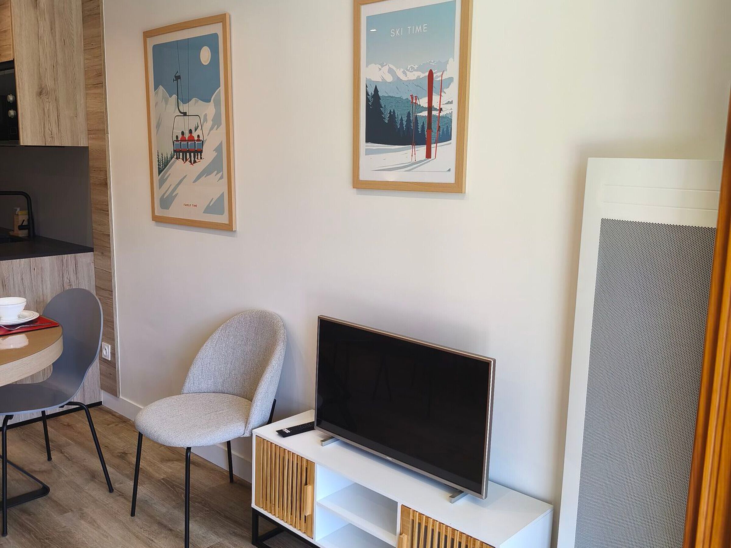 A modern living room with a flat-screen TV, a grey upholstered chair, a small table with a bowl, and two framed pictures on the wall.