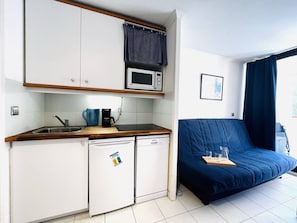 A compact kitchenette with a microwave, coffee maker, and dishwasher. A blue sofa is positioned next to the kitchen, and a window with curtains is visible. - Studio with direct beach access - 4 guests (Le Lavandou)
