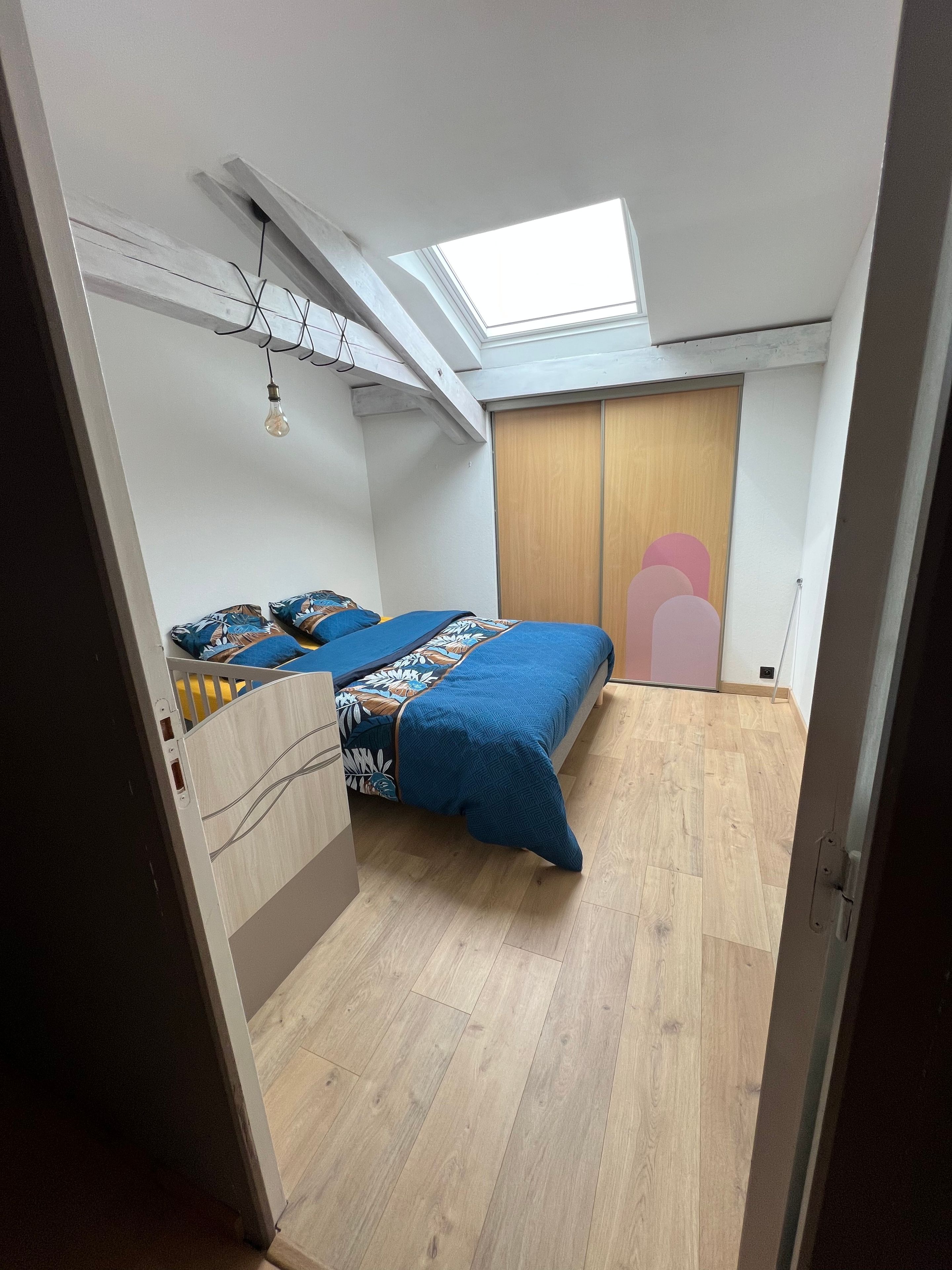 Une chambre avec un puits de lumière, un lit recouvert d’une couverture bleue et une armoire.
