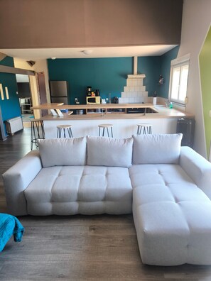 A modern living room with a sectional sofa, a kitchen area with a microwave and coffee maker, and a dining area with stools.