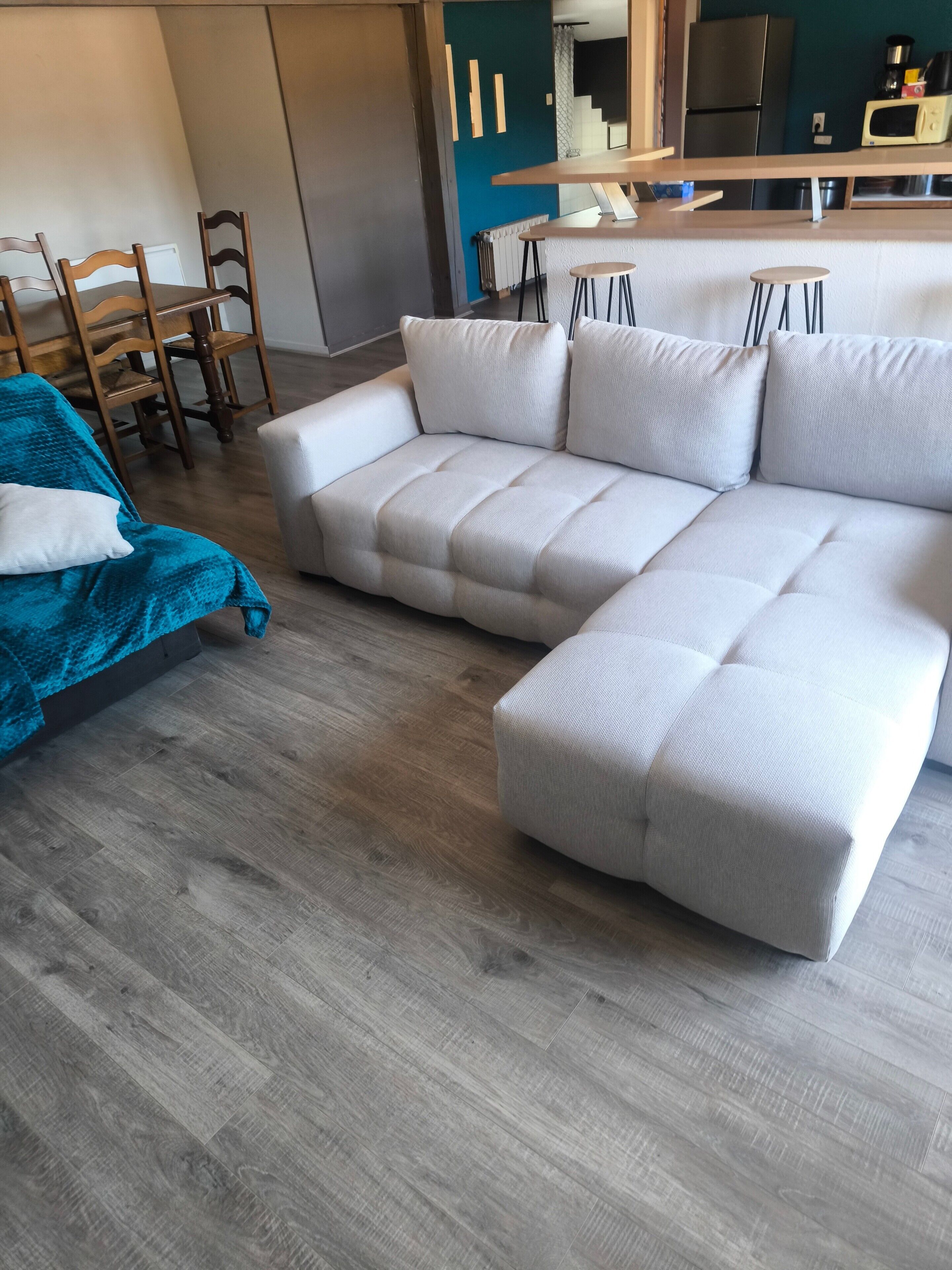 A modern living room with a sectional sofa, wooden chairs, a blue blanket, and a kitchen area in the background.