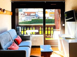 A room with a sofa, a small table, a TV, and a view of a building through a glass door. - Bright duplex near center - 6 guests (Les Gets)