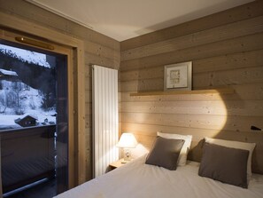 A bedroom with a wooden wall, a bed with pillows, a nightstand with a lamp, and a window with a view of a snowy landscape. - Refined apartment in Méribel center - 6 guests (Méribel)
