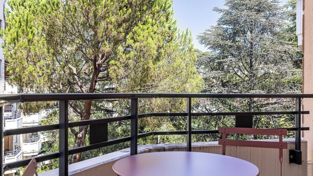 Un balcón con una mesa redonda y una silla, con vista a un follaje denso y un edificio.