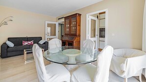 Una sala moderna con una mesa de comedor de vidrio rodeada de sillas blancas, un sofá gris oscuro con cojines estampados y un aparador de madera con puertas de cristal.