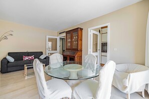 Ein modernes Wohnzimmer mit einem Esstisch aus Glas, umgeben von weißen Stühlen, einem dunkelgrauen Sofa mit gemusterten Kissen und einer Holzkabine mit Glastüren.