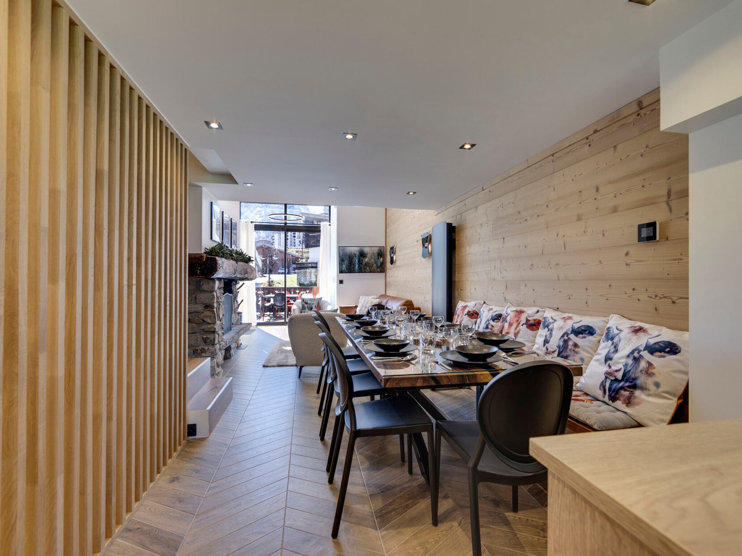 A modern dining area with a long table set for multiple people, wooden paneling on the walls, and a view into the adjacent room.