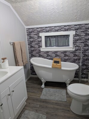 Un baño con una bañera independiente, inodoro, tocador con lavamanos y ventana con cortina.