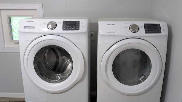 Deux machines à laver Samsung blanches se trouvent dans une pièce dont le sol est en bois et qui possède une fenêtre donnant sur un paysage verdoyant.