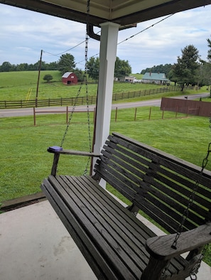 Een houten bank met een metalen schommel, een witte kolom en uitzicht op een landelijk landschap met een schuur en een weg.