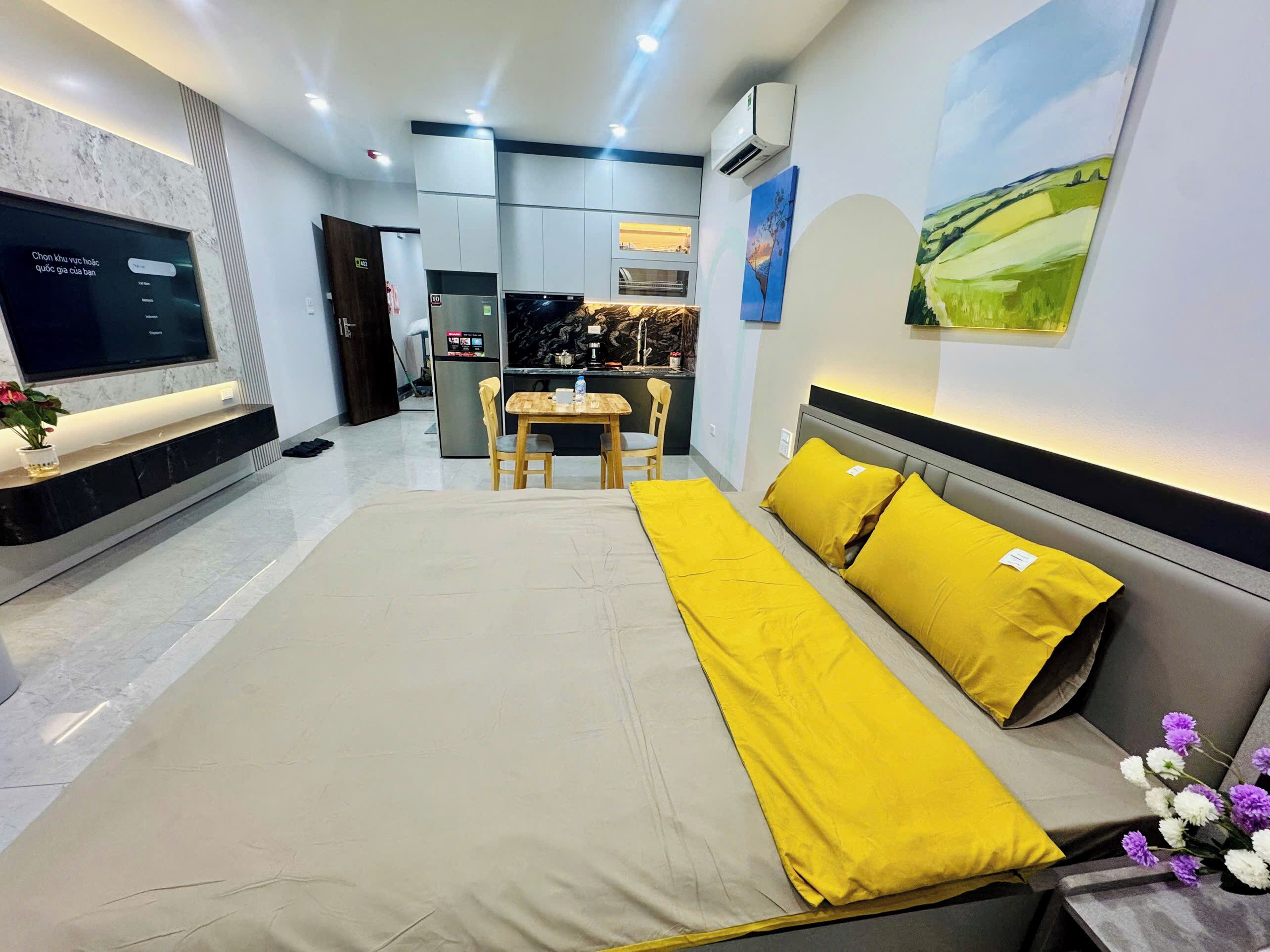 A modern hotel room with a large bed, a TV, a dining area, and a kitchen in the background.