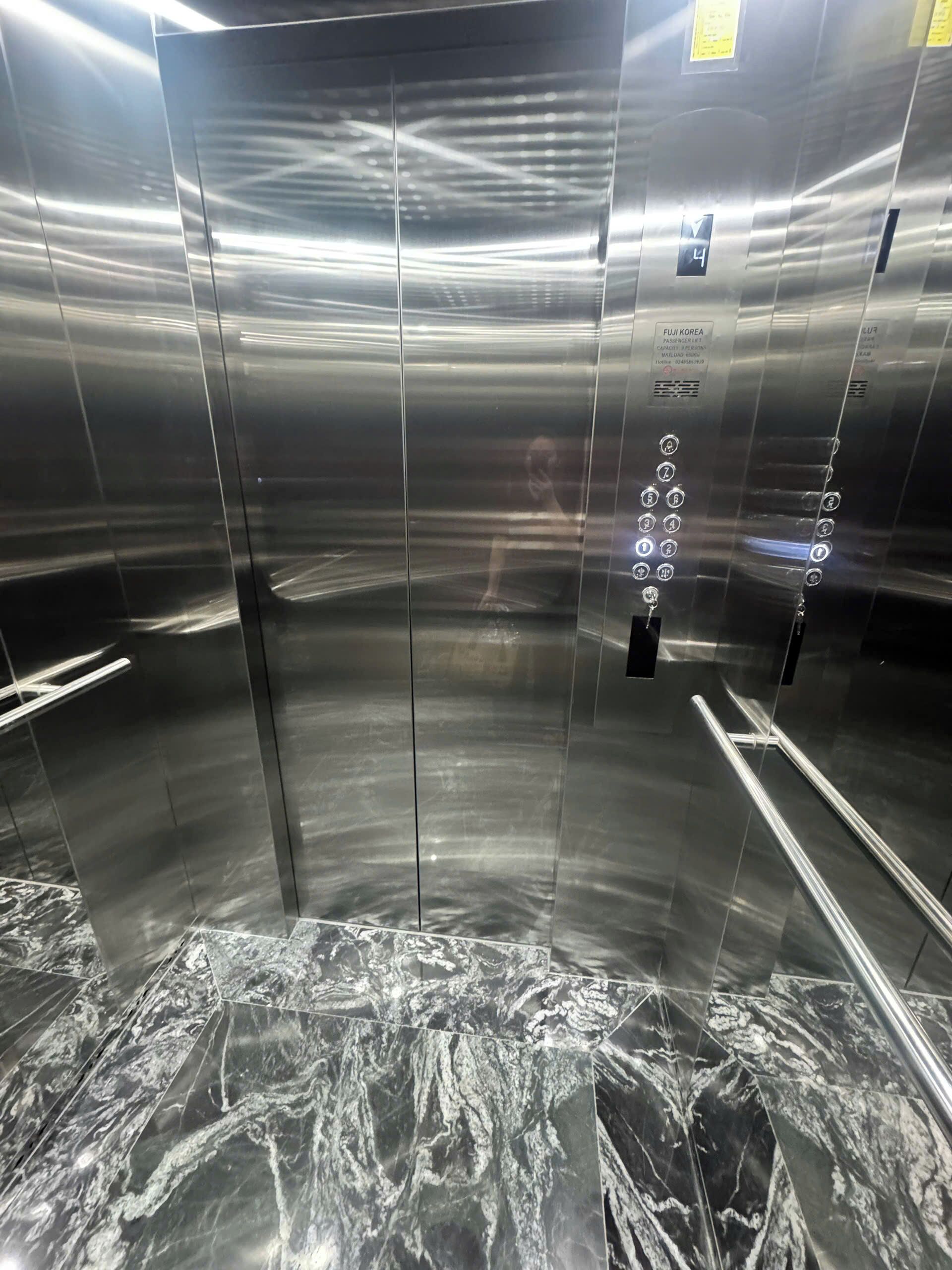 Elevatorinteriør med vægge i metal, en marmorgulv og en person synlig gennem glasset.