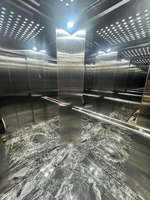 An elevator interior with metallic walls, a marble floor, and overhead lighting.