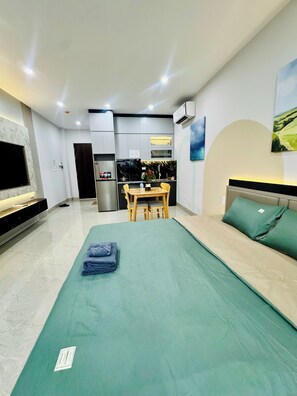 A modern hotel room with a bed, a dining table with chairs, a kitchen area, and a TV mounted on the wall.