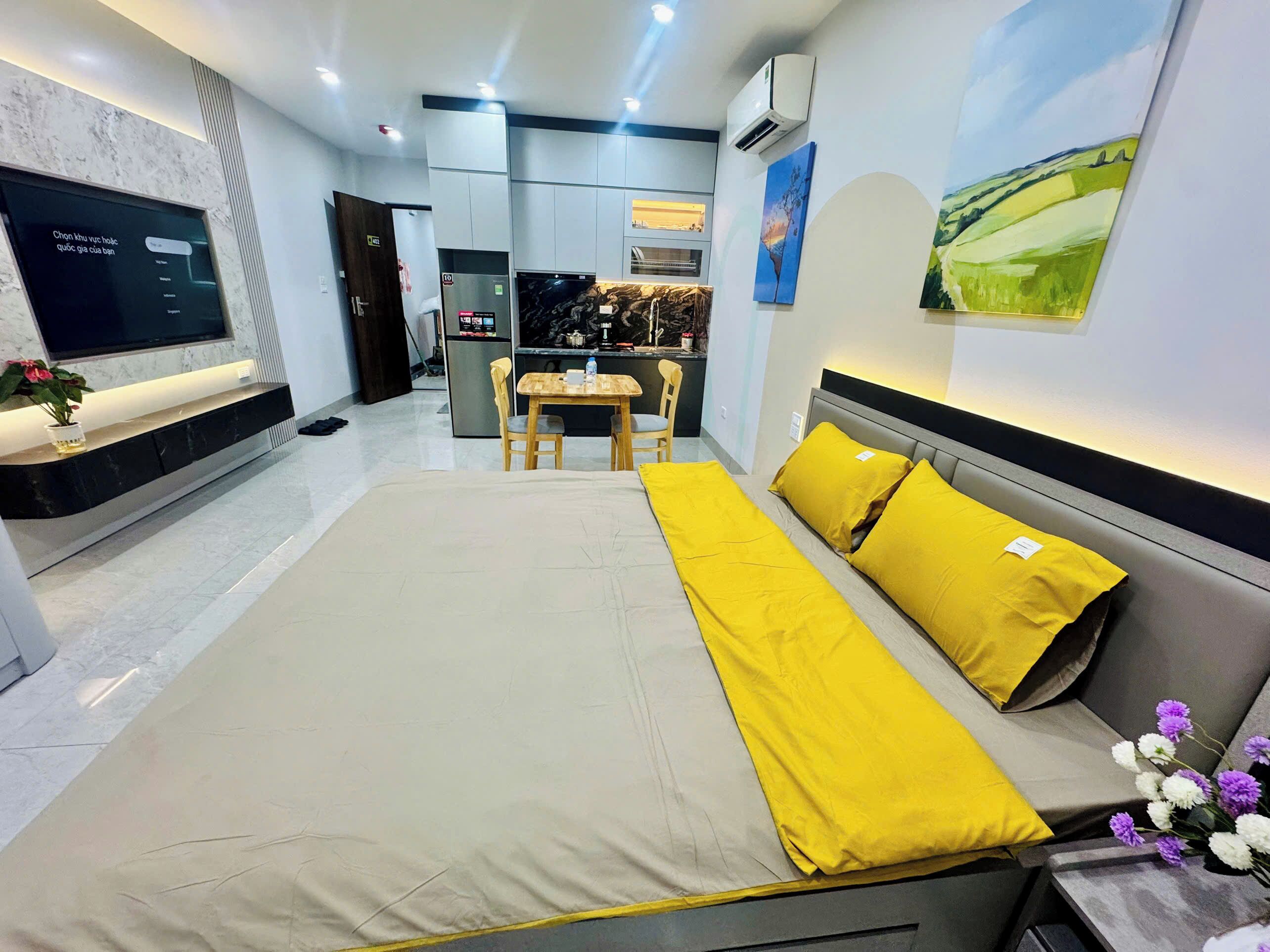A modern hotel room with a bed, a desk, and a kitchenette in the background.