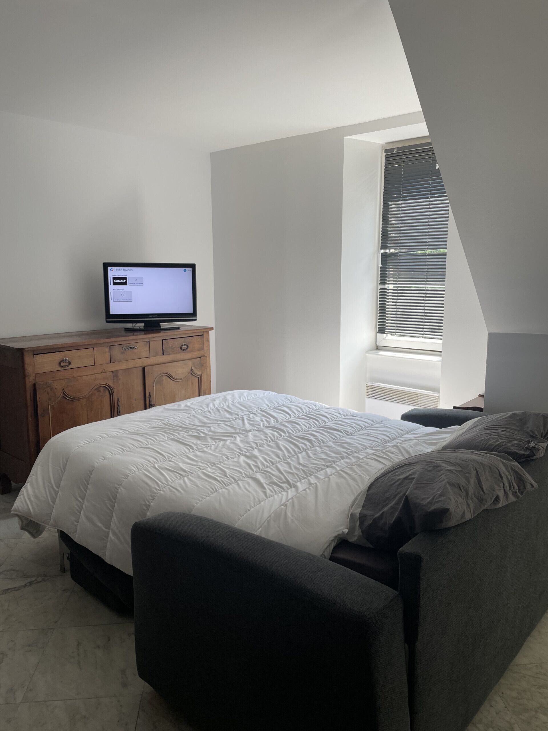 Een slaapkamer met een bed, een televisie op een houten dressoir en een raam met jaloezieën.