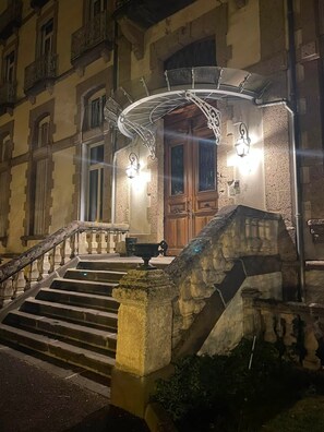 A building with a stone staircase leading to a wooden door, a metal canopy, and two wall-mounted lights. - APPARTEMENT DUPLEX RESIDENCE DU PARC (Saint-Nectaire)