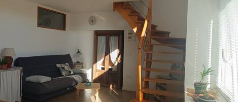 Una sala de estar con un sofá, una mesita, una escalera de madera y una ventana con persianas.