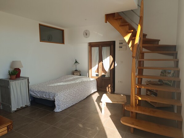 A bedroom with a bed, a nightstand, a lamp, a window with curtains, a wooden staircase, and a wall clock.